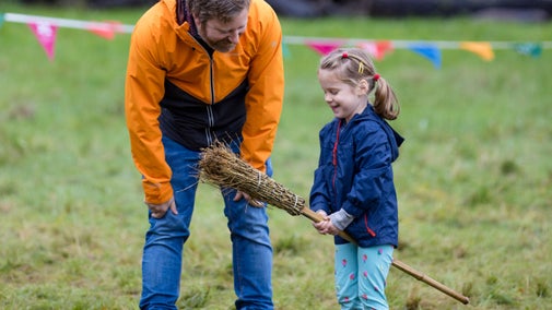 Young girl in a bright coloured patterned jacket pretending to fly on a Broomstick with her dad cheering her on